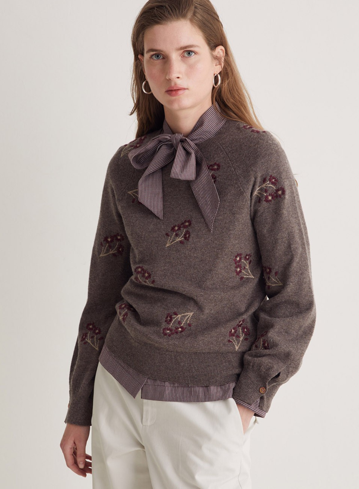 Woman wearing a brown sweater with floral embellishments and a bow tie on a plain background