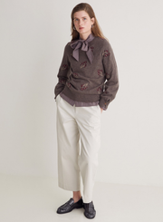 Woman wearing a brown embroidered sweater with a bow tie, white pants, and black shoes on a light gray background