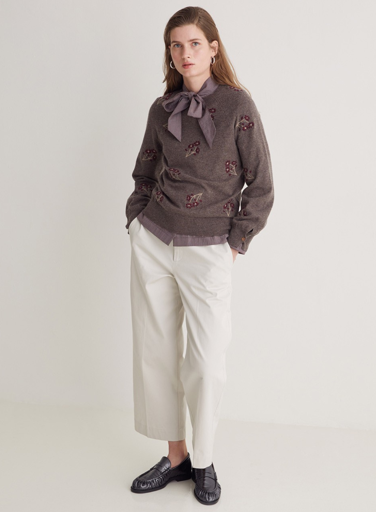 Woman wearing a brown embroidered sweater with a bow tie, white pants, and black shoes on a light gray background