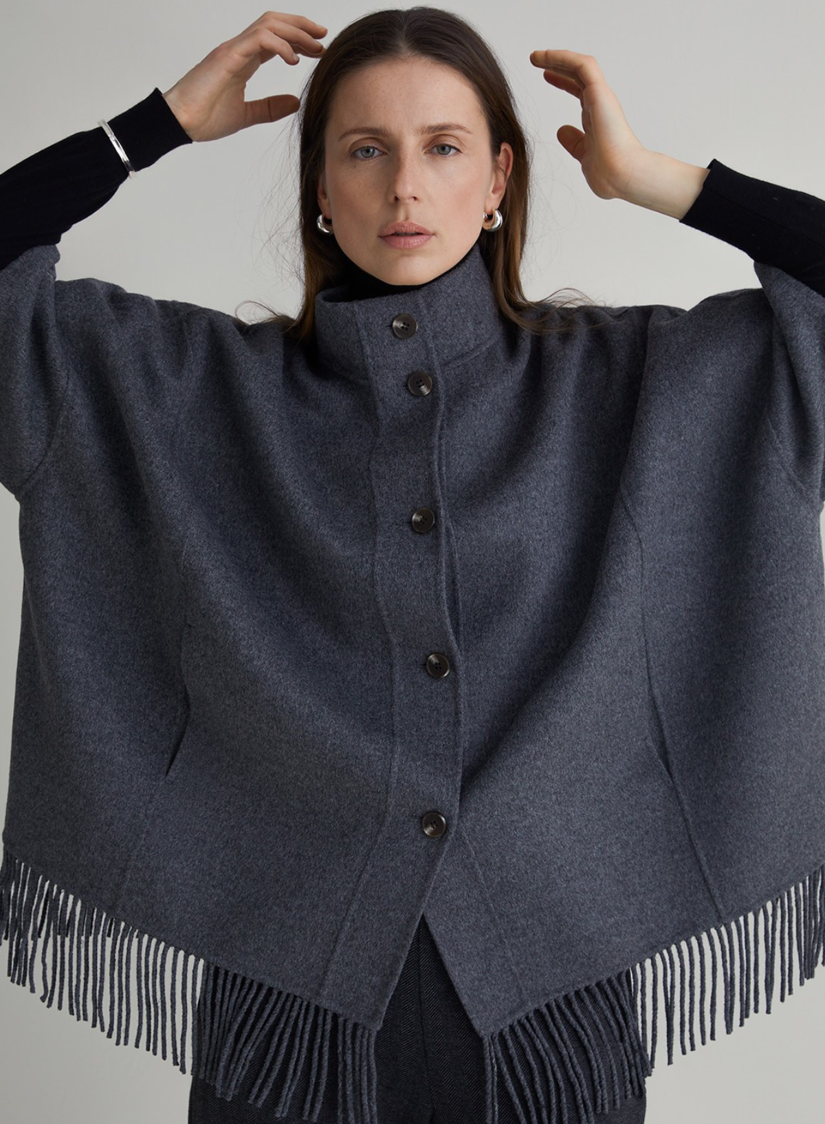 Woman wearing a dark gray poncho with fringe details on a plain background