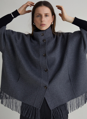 Woman wearing a dark gray poncho with fringe details on a plain background