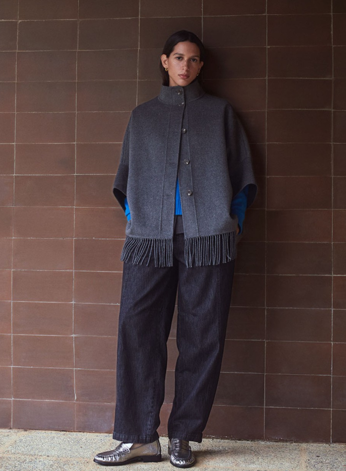 Person wearing a gray poncho with fringe standing against a brown tiled wall.