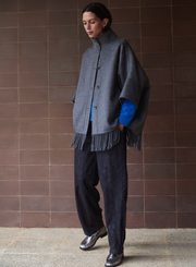 Person wearing a gray poncho with fringe, black trousers, and blue shirt against a brown tiled wall.