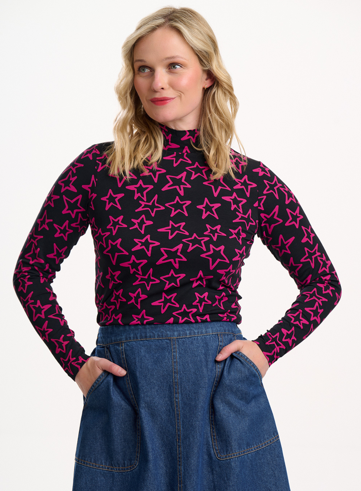 Woman wearing the Sugarhill Brighton Annika Black Pink Doodle Stars Turtleneck Jersey Top and a blue denim skirt against a white background.