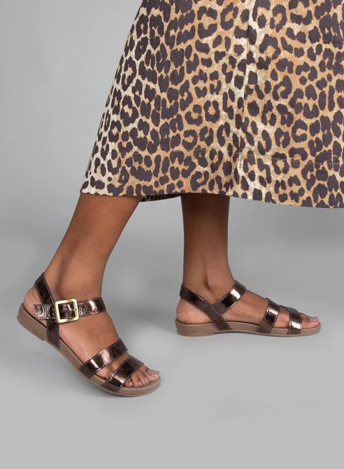 Model wearing the Esska Ilona Metallic Coffee Flat Sandals on a grey backdrop styled with a leopard print dress. Model is in side profile showcasing the metallic straps.