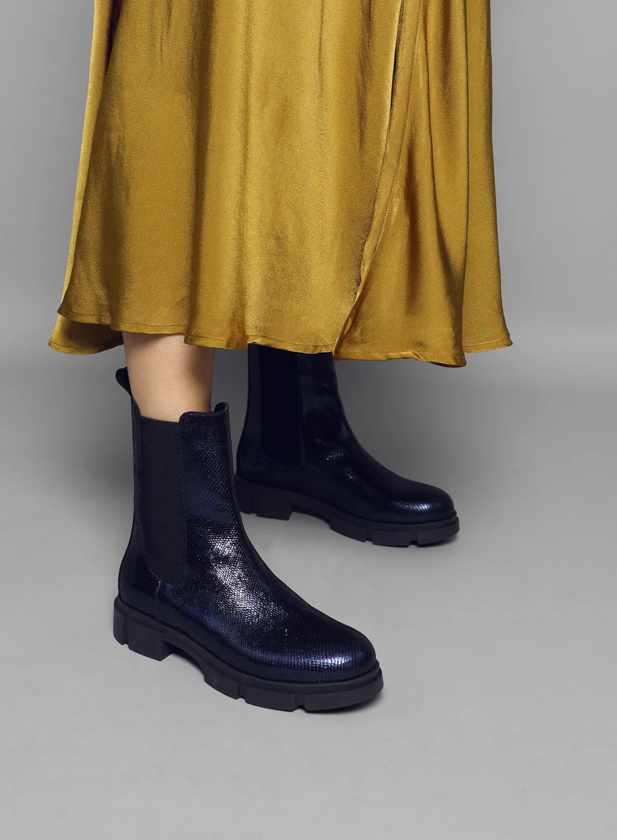 Model wearing an ochre yellow satin skirt styled with Esska June Navy Metallic Chelsea Boots on a grey background. Model is in side front profile showcasing the round toe shape and the metallic appearance of the Chelsea boots.