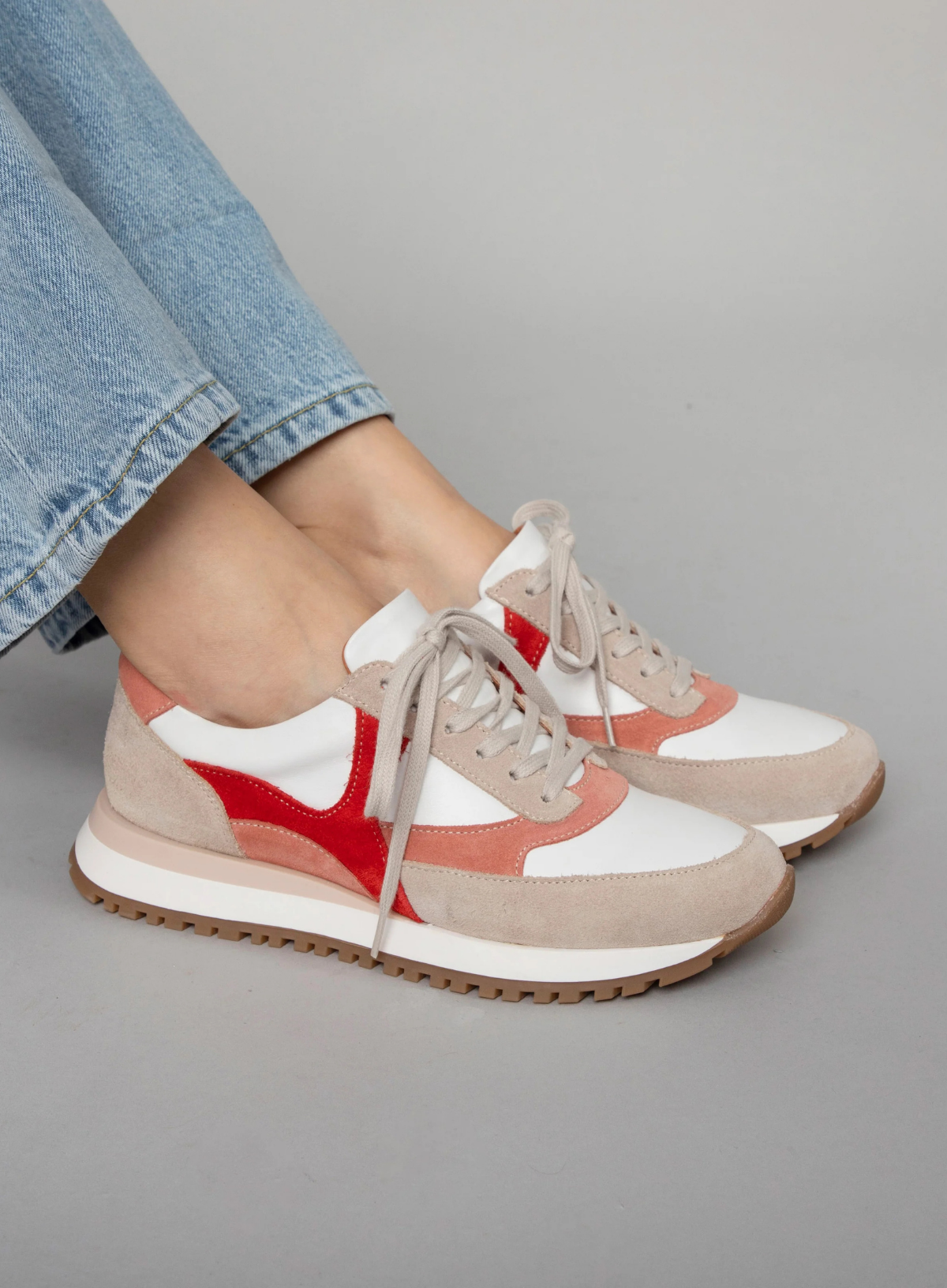Model wearing the Esska Cinda White and Pink Suede Trainers with light wash denim jeans on a grey backdrop. Side shot showcasing the round toe shape of the trainers and the suede detail.