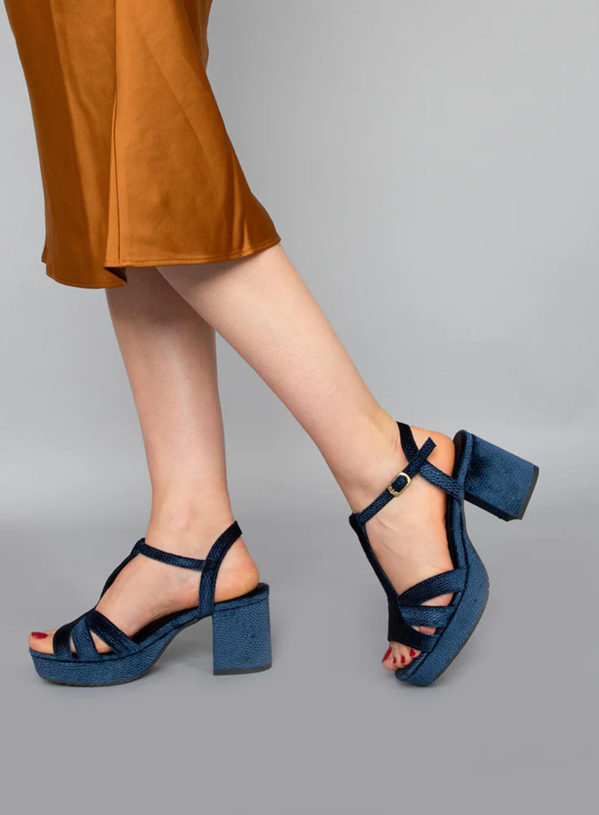 Model wearing the Esska Charlie Blue Velvet T Bar Heels on a grey background. Model has styled the block heels with a burnt orange satin skirt and is in side profile showcasing the breathable design.