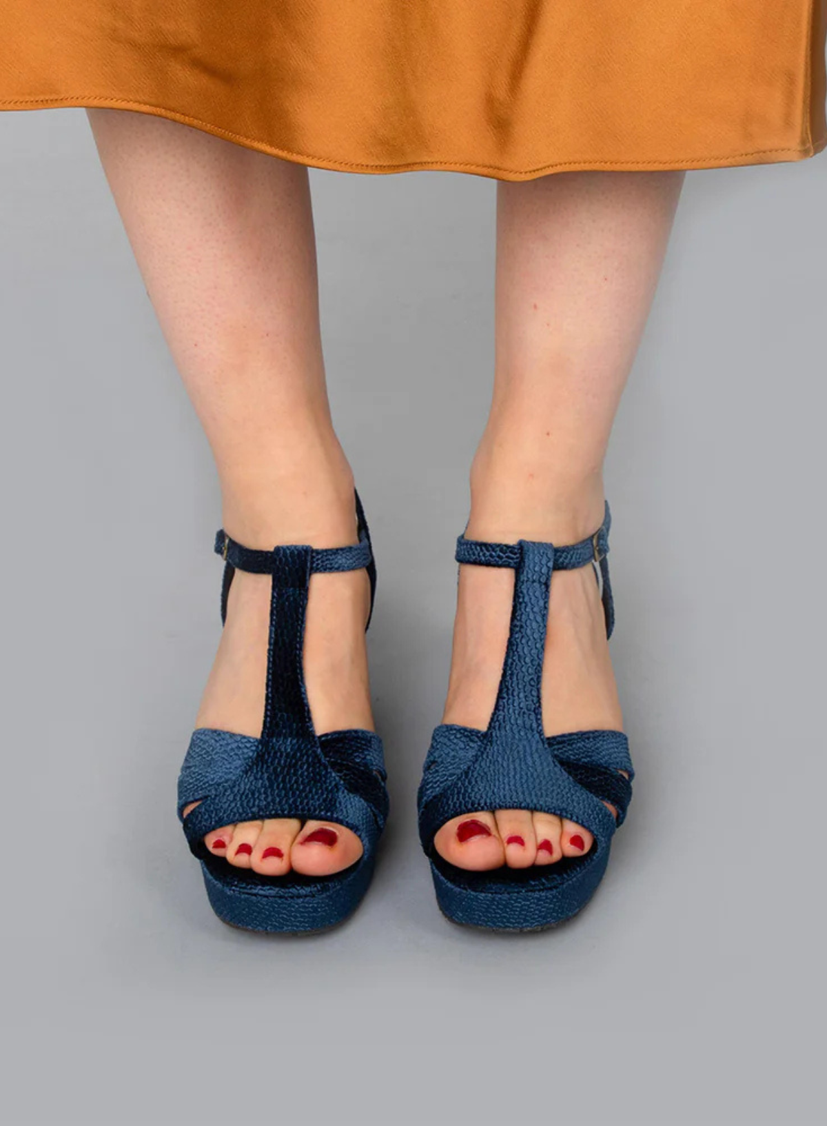 Model wearing the Esska Charlie Blue Velvet T Bar Heels on a grey backdrop styled with a burnt orange satin skirt. Front shot showcasing the open toe design and the t bar straps.