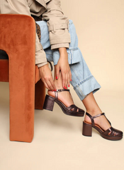 Model styling the Esska Celeste Dark Brown Metallic Platform Block Heels with a pair of light wash jeans, rolled up with a beige Mac jacket and camel colour ribbed jumper. Model is sitting on an orange chair adjusting the heel straps while showcasing the block heels.