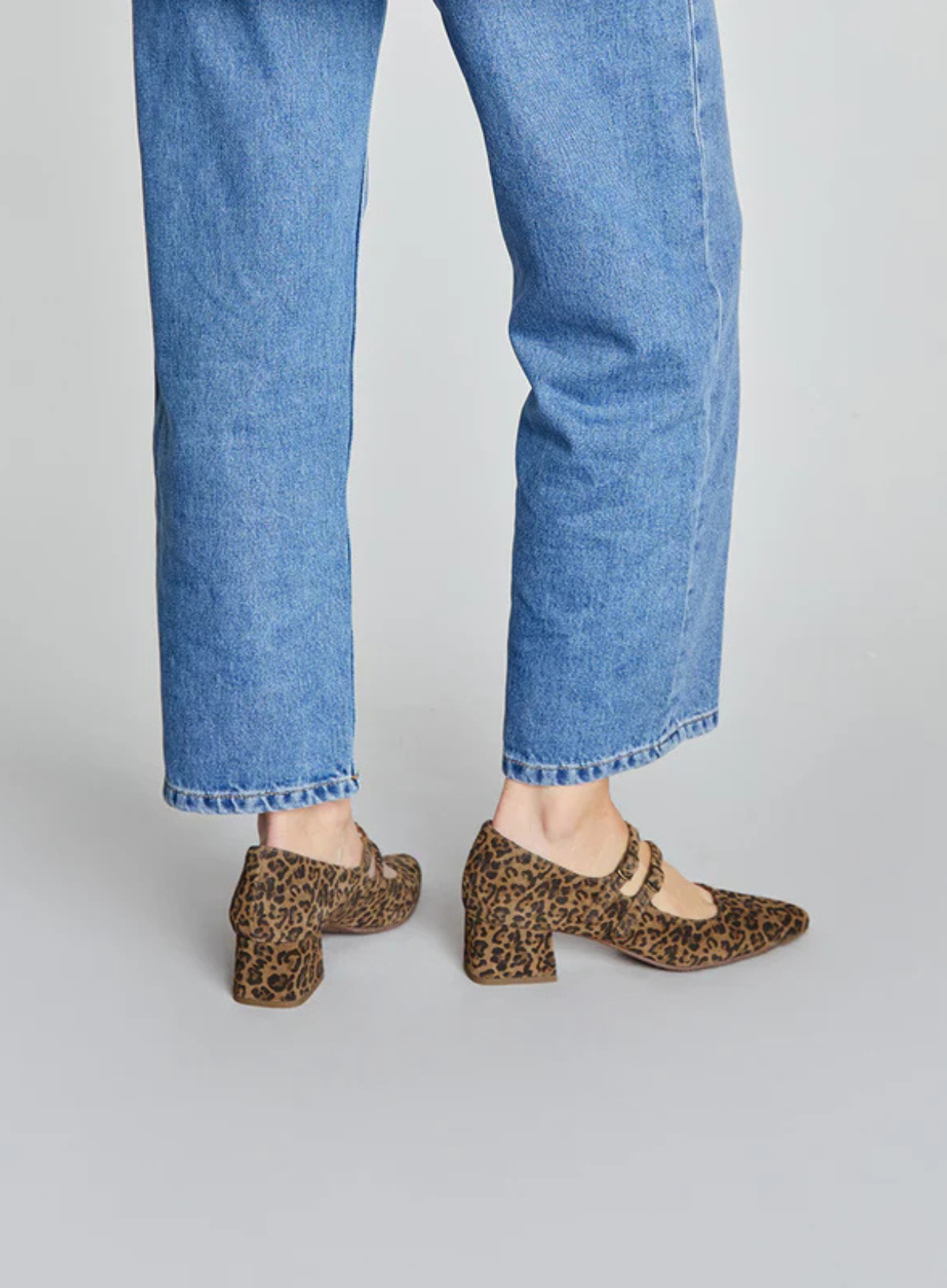 Woman wearing the Esska Laura Low Leopard Heels and blue jeans on a plain background. Back shot showcasing the block heel on these Mary Janes.