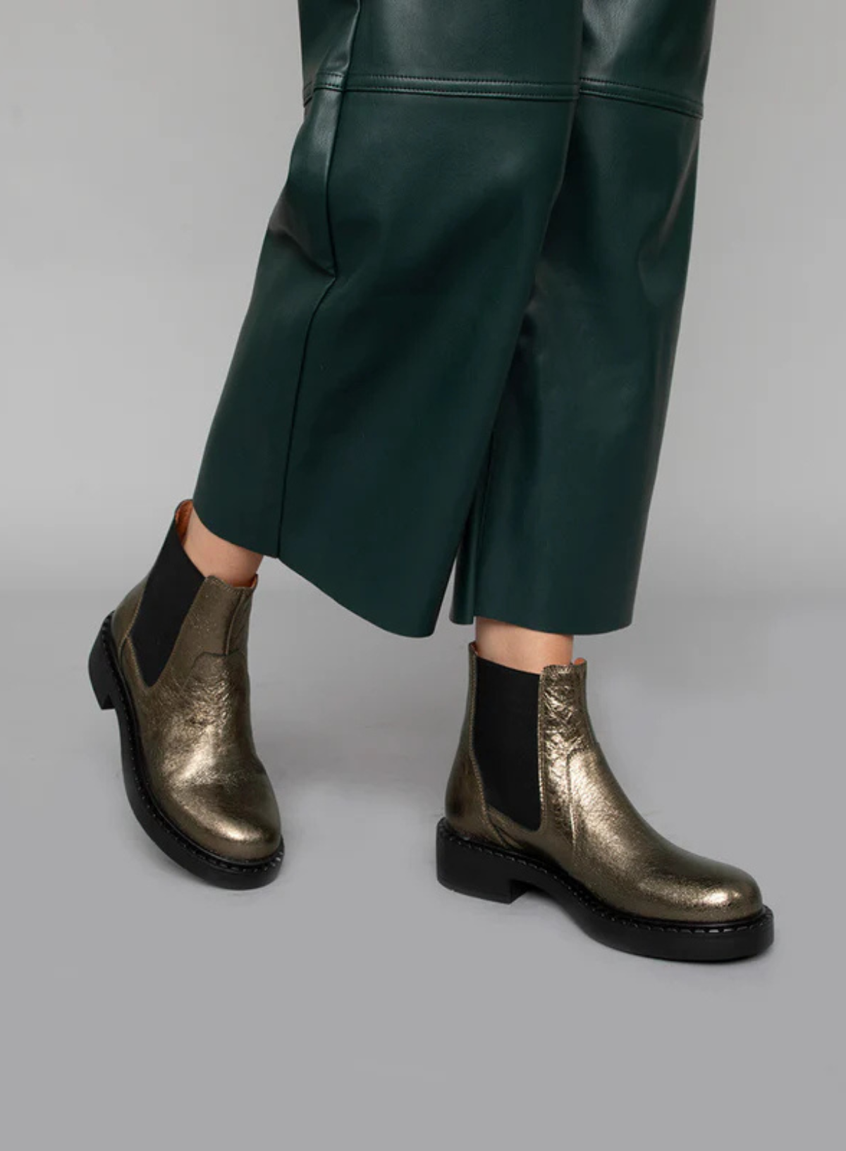 Model wearing the Esska Bora Graphito Leather Chelsea Boots styled with green leather wide leg trousers on a gray background.