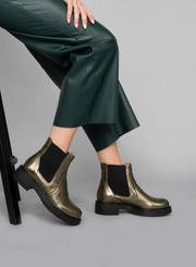Model wearing green leather wide leg trousers with the Esska Bora Graphito Leather Chelsea Boots sitting on a stool in side profile. Model is showcasing the block heel to these leather boots.