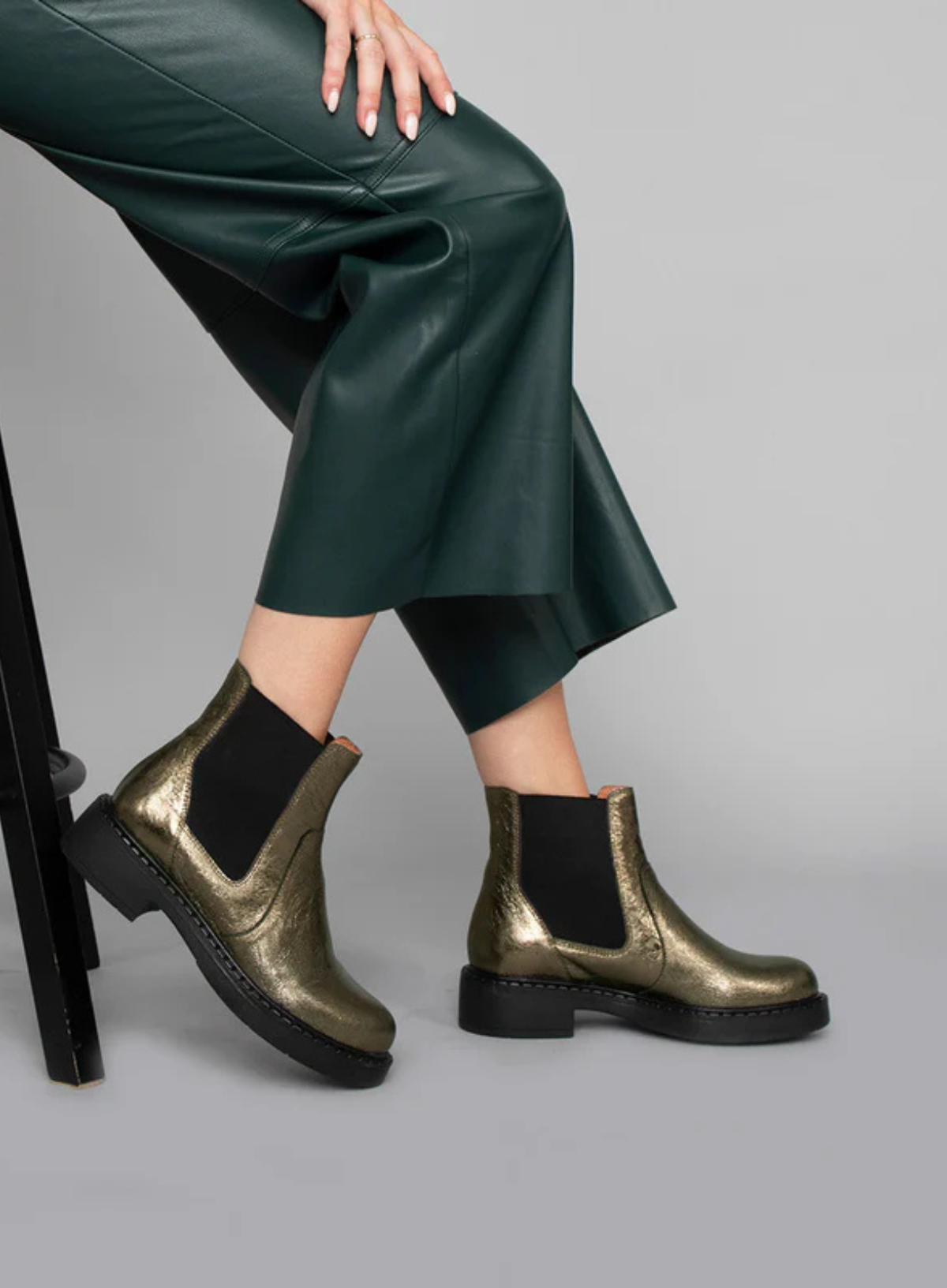Model wearing green leather wide leg trousers with the Esska Bora Graphito Leather Chelsea Boots sitting on a stool in side profile. Model is showcasing the block heel to these leather boots.