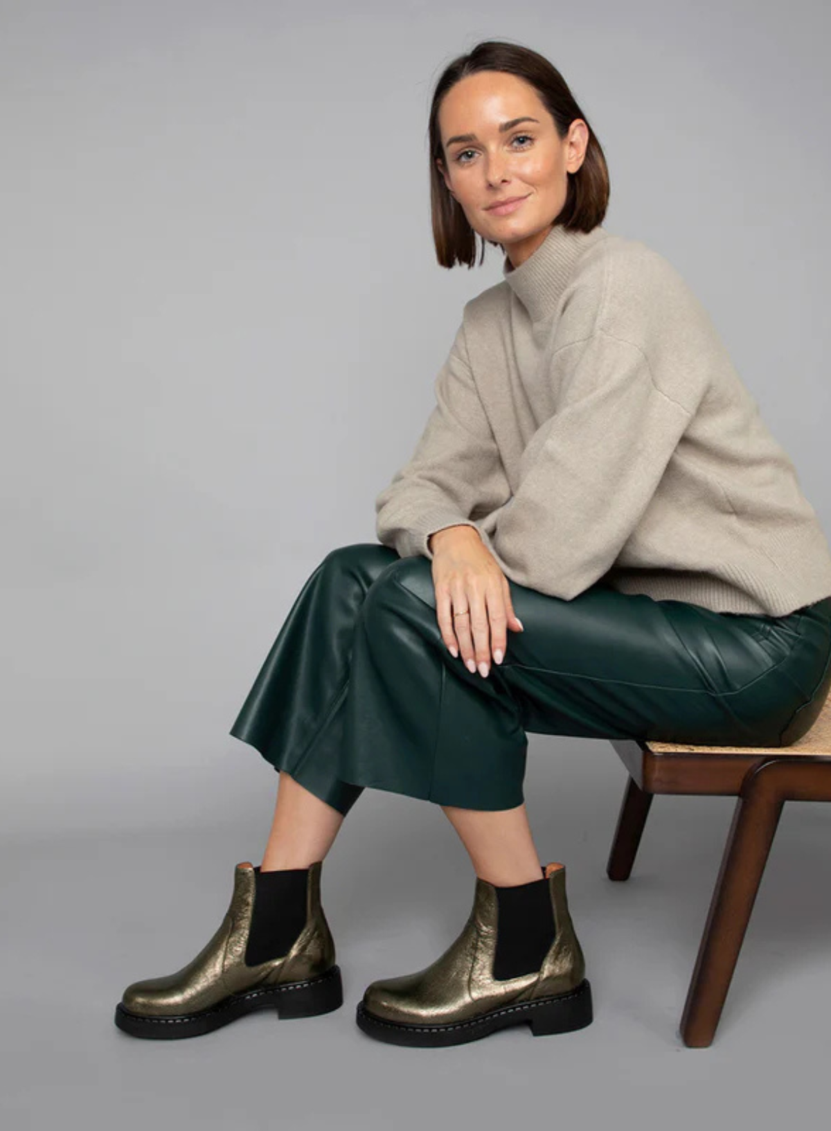 Model sitting on a chair wearing a beige sweater, green wide leg leather trousers styled with the Esska Bora Graphito Leather Chelsea Boots against a grey background.
