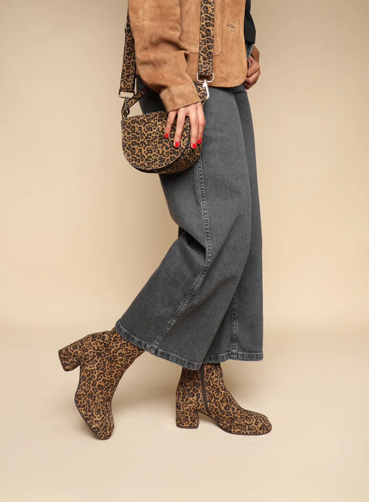 Model wearing the Esska Mona Leopard Print Boots, holding a matching handbag, with a tan suede jacket and charcoal wide leg jeans on in side profile, showcasing how the ankle boots can be styled.