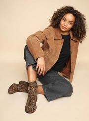 Woman wearing a brown suede jacket a black top and charcoal jeans with the Esska Mona Leopard Print Boots on a beige background showcasing how the ankle boots can be styled.