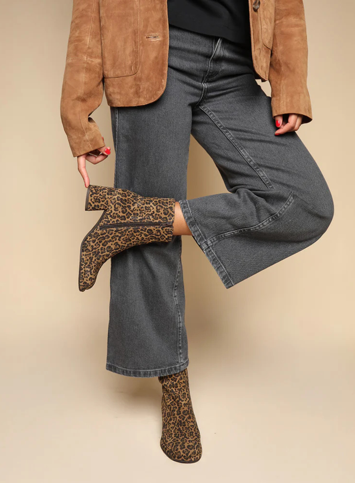 Model wearing the Esska Mona Leopard Print Boots a brown suede jacket, black top and charcoal wide leg jeans on a beige background. The model is showcasing the block heel and zip closure of these ankle boots.