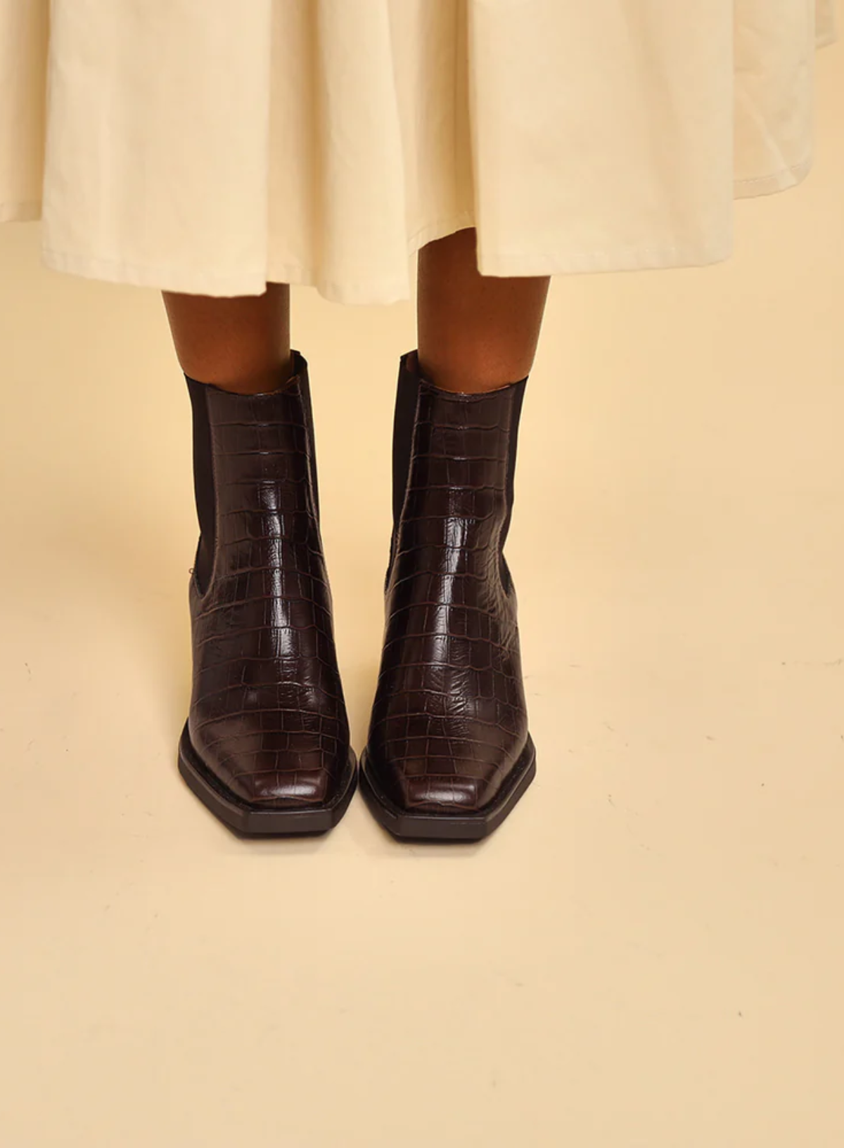 Model wearing the Esska Gillian Dark Brown Embossed Croc Ankle Boots with a cream skirt. Close up front shot showcasing the square toe shape and the embossed crocodile pattern to these Chelsea boots.