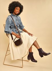 Woman sitting on a chair wearing a denim shirt, white skirt, and the Esska Gillian Dark Brown Embossed Croc Ankle Boots, holding a brown handbag showcasing how they can be styled.
