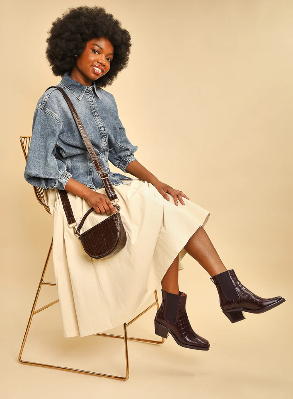 Woman sitting on a chair wearing a denim shirt, white skirt, and the Esska Gillian Dark Brown Embossed Croc Ankle Boots, holding a brown handbag showcasing how they can be styled.