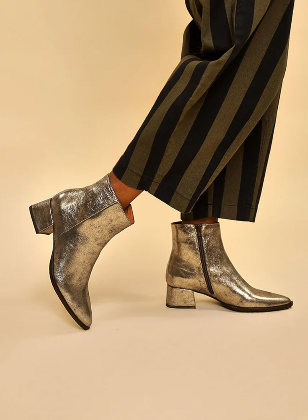 Esska Luka Gold Ankle Boots worn with striped trousers on a beige background. Side shot showcasing block heel and zip closure.
