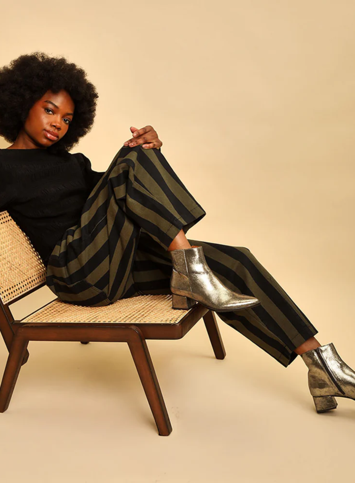 Model shot wearing the Esska Luka Gold Ankle Boots with green and black trousers on with a black jumper sitting on a low chair.