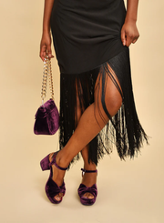 Person wearing a black fringe dress with the Esska Chilli Aubergine Velvet Heels and a matching handbag on a beige background.