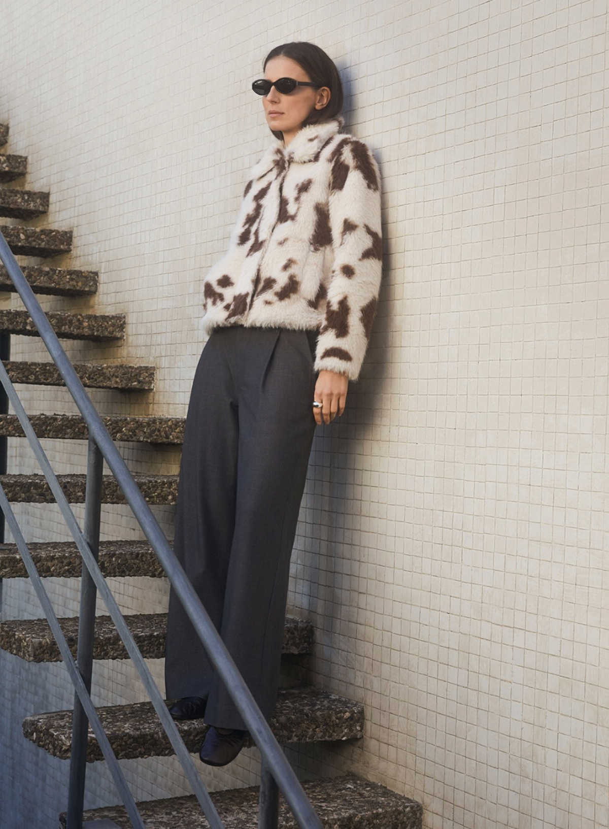 Person wearing a patterned coat leaning against a tiled wall next to stairs.
