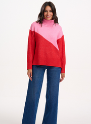 Woman wearing the Sugarhill Brighton Winona Pink Red Colour Block Roll Neck Jumper with blue jeans on a white background