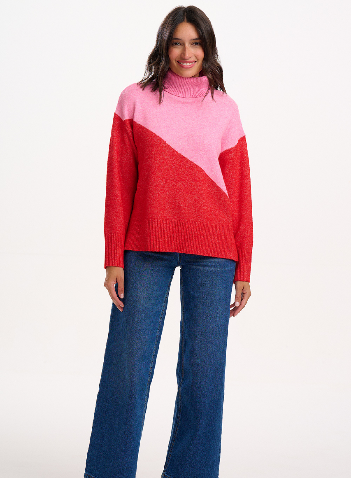Woman wearing the Sugarhill Brighton Winona Pink Red Colour Block Roll Neck Jumper with blue jeans on a white background