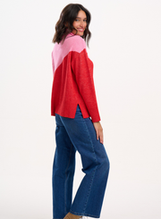 Woman wearing the Sugarhill Brighton Winona Pink Red Colour Block Roll Neck Jumper with blue jeans on a white background side profile showcasing jumper hem slit
