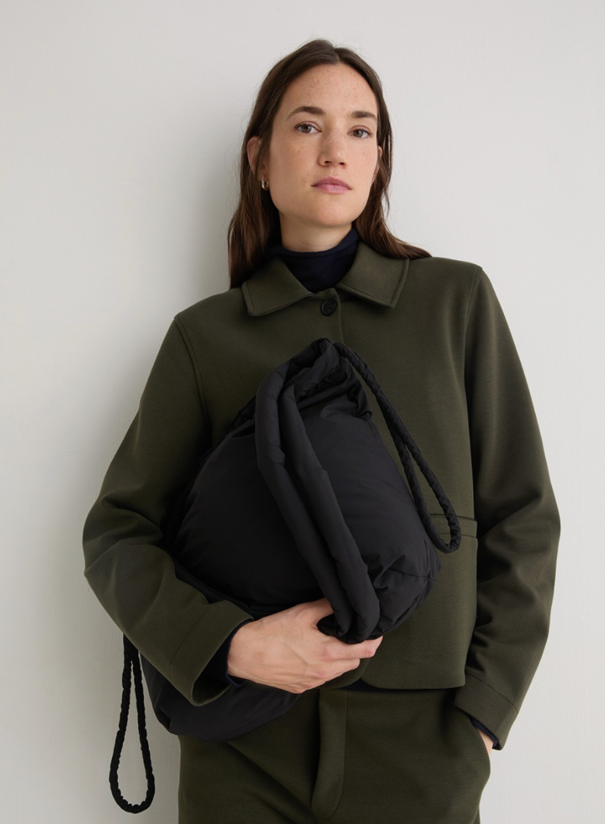 Person wearing a green coat holding a black bag against a plain background