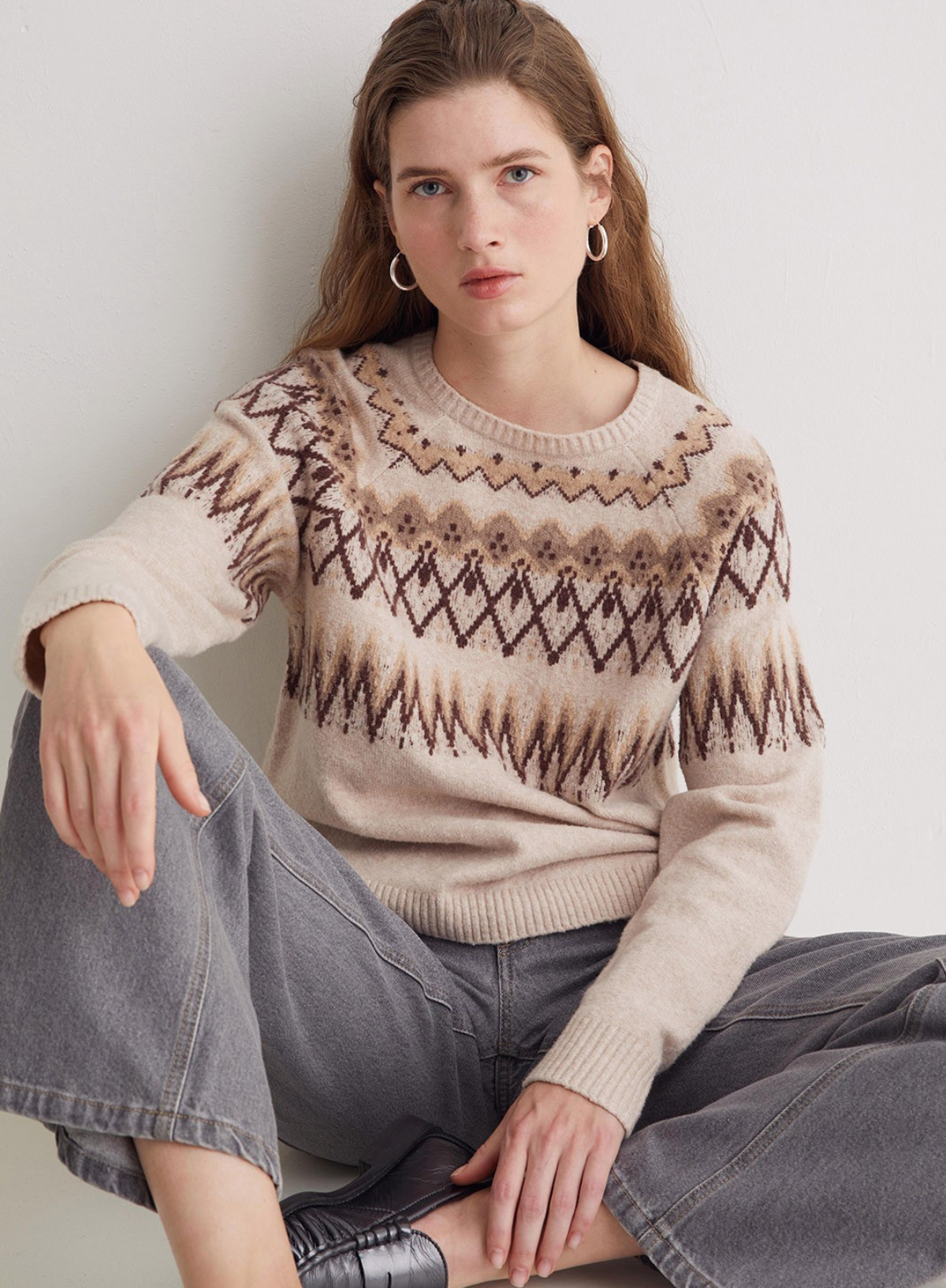 Woman wearing a patterned sweater and jeans sitting against a plain background